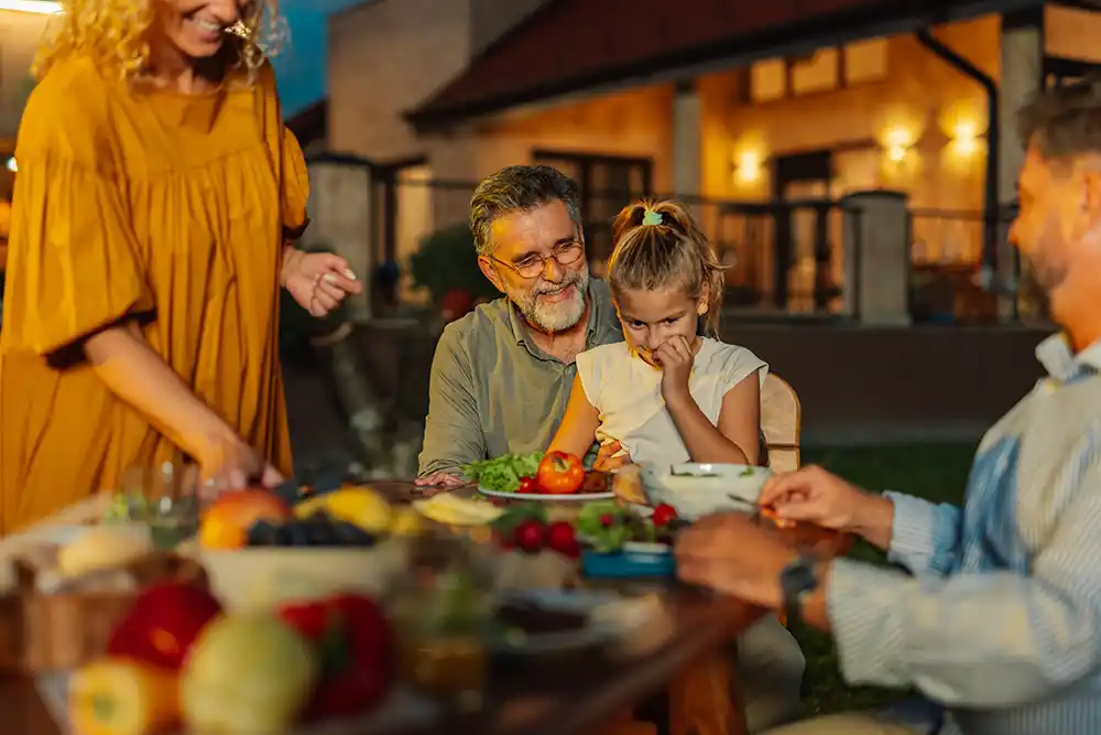Family gathered around an outdoor dinner table in the evening, with a smiling bearded man, a young girl, and a lit house behind.