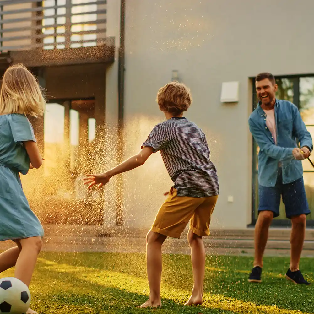 Children and a father playing with a spraying garden hose on a backyard lawn at sunset, a soccer ball on the grass.