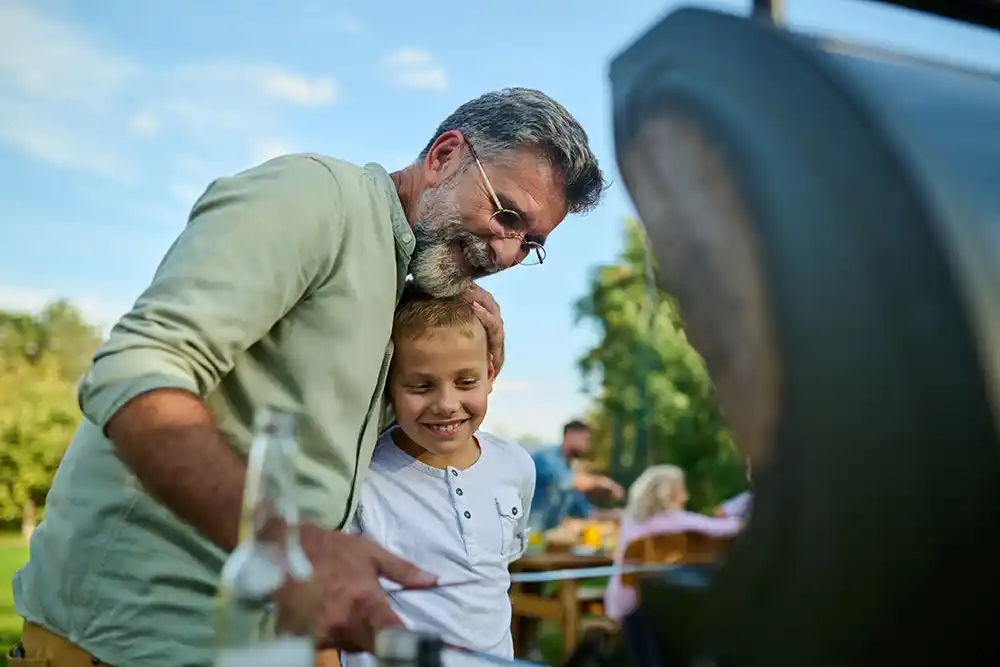 Man with glasses embracing a smiling young boy beside a barbecue grill outdoors, with family seated at a table behind.